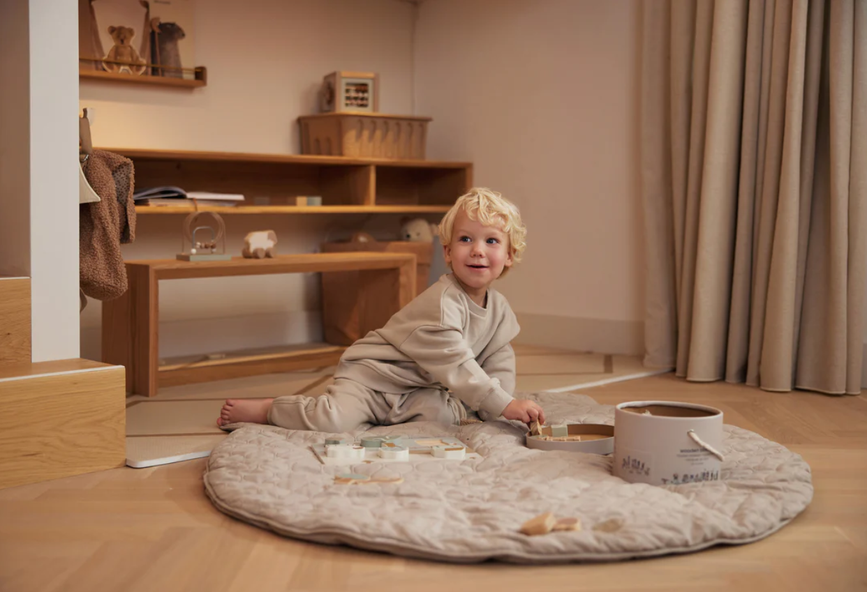 Tapete acolchoado redondo cinza claro com menino loiro a brincar em cima, em interior com móveis de madeira