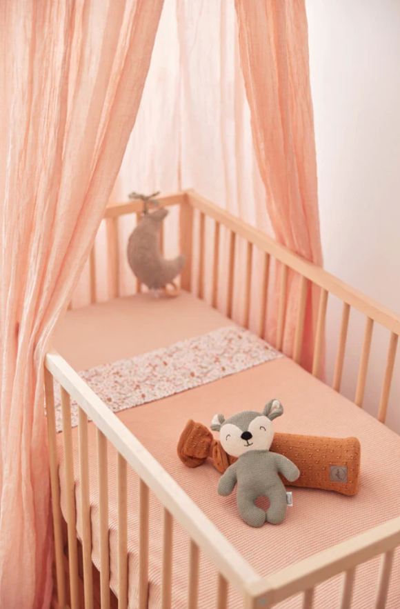 Berço de madeira clara com colchão cor de salmão e cortinas rosa, brinquedos de pelúcia em cima