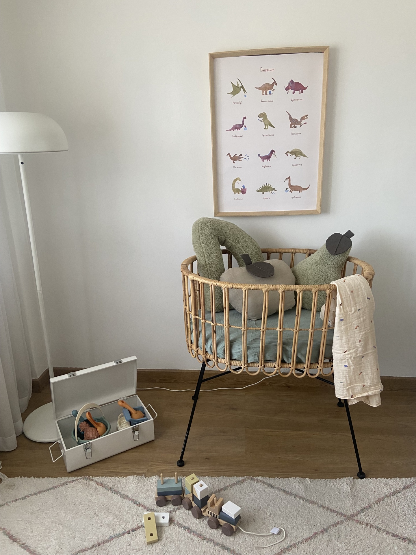 Berço de madeira clara com almofadas verdes e bege, candeeiro branco, caixa com brinquedos e poster de dinossauros na parede.