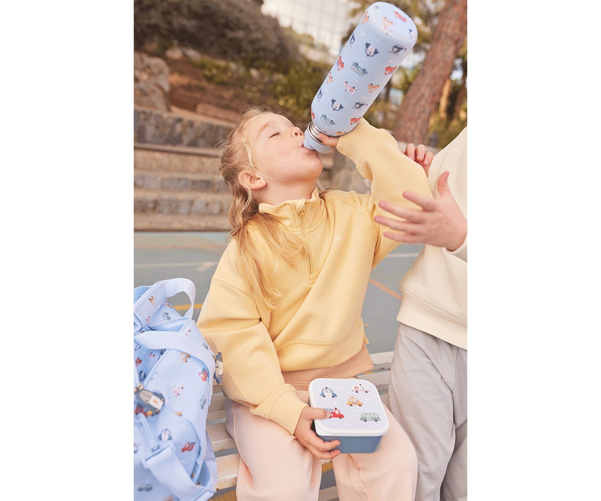 Menina a beber água de garrafa azul com padrão, segurando lancheira azul, com mochila azul ao lado