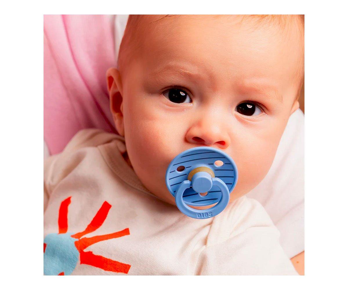 Bebé com chupeta azul clara em camisola branca com padrão vermelho e azul