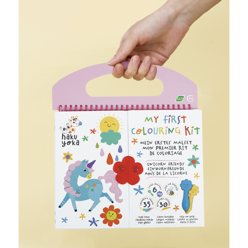 Livro de colorir infantil com unicórnio e flores na capa, embalagem com alça rosa e uma mão a segurar.