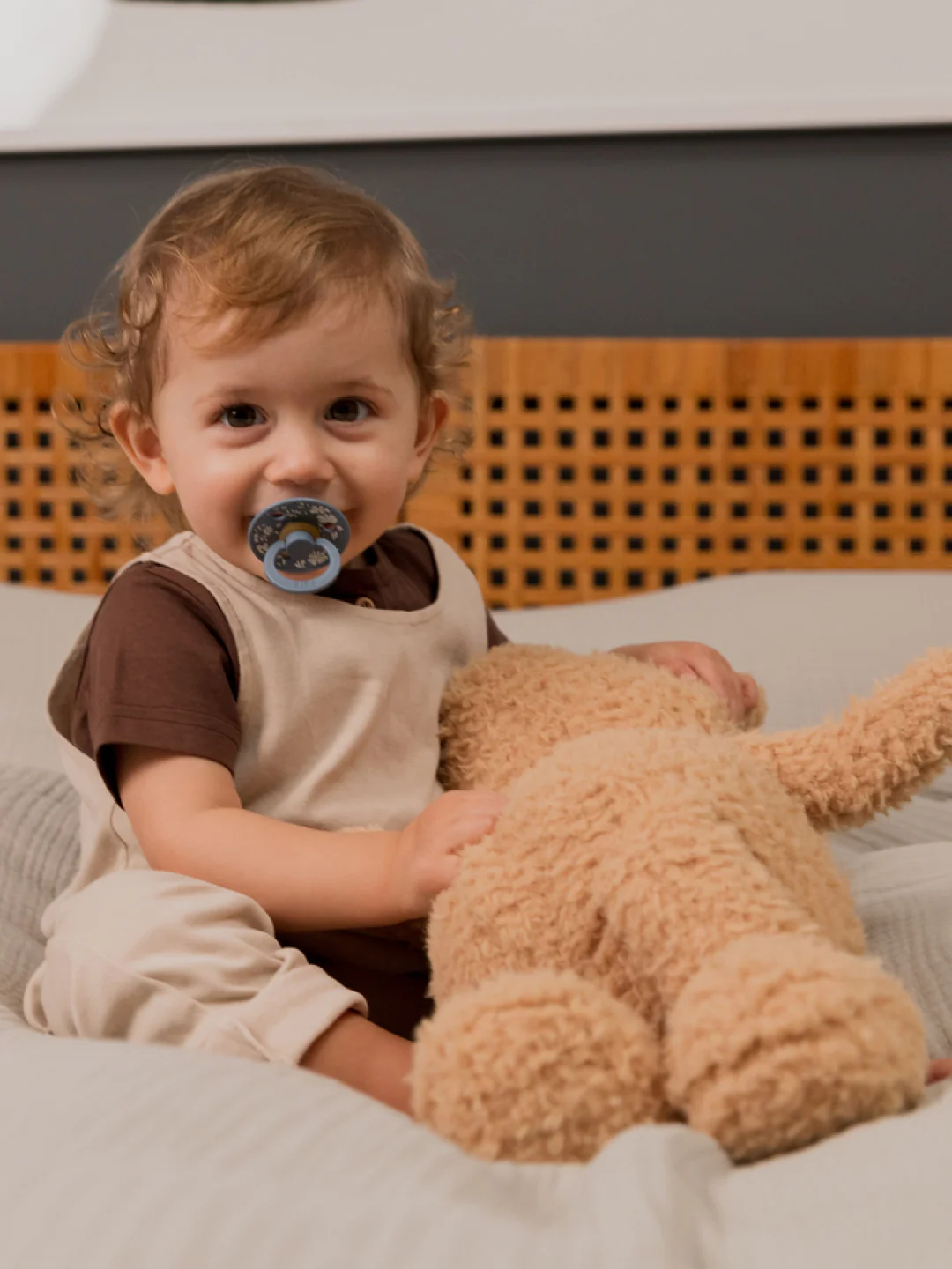 Criança com babygrow bege e chupeta cinzenta segurando urso de peluche numa cama