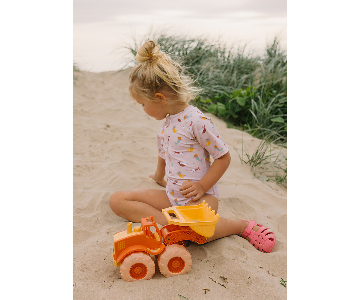 Criança com fato de banho rosa e trator de brinquedo na areia com vegetação