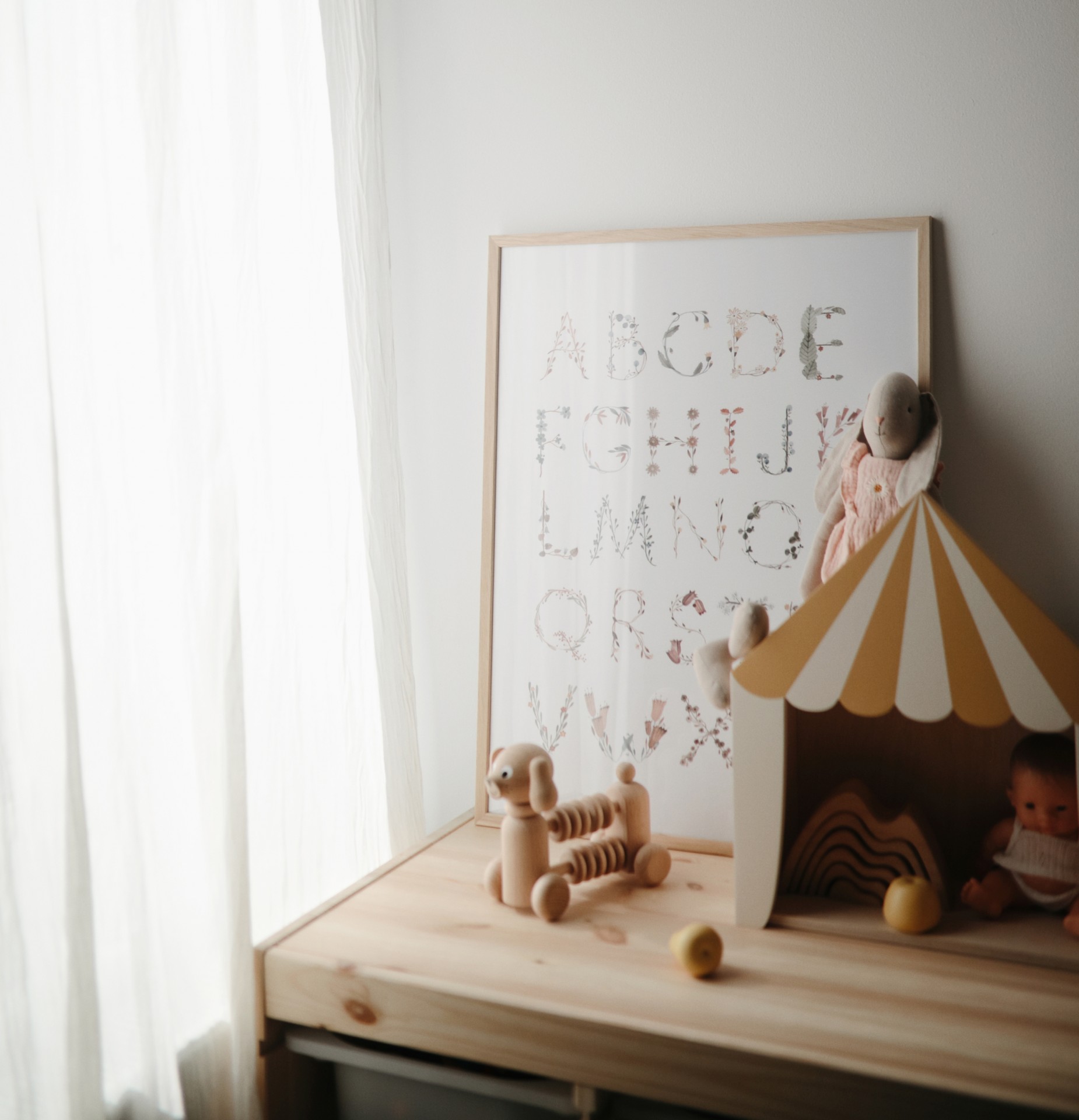 Brinquedos de madeira e quadro de alfabeto num móvel de madeira clara perto de uma janela com cortina branca.