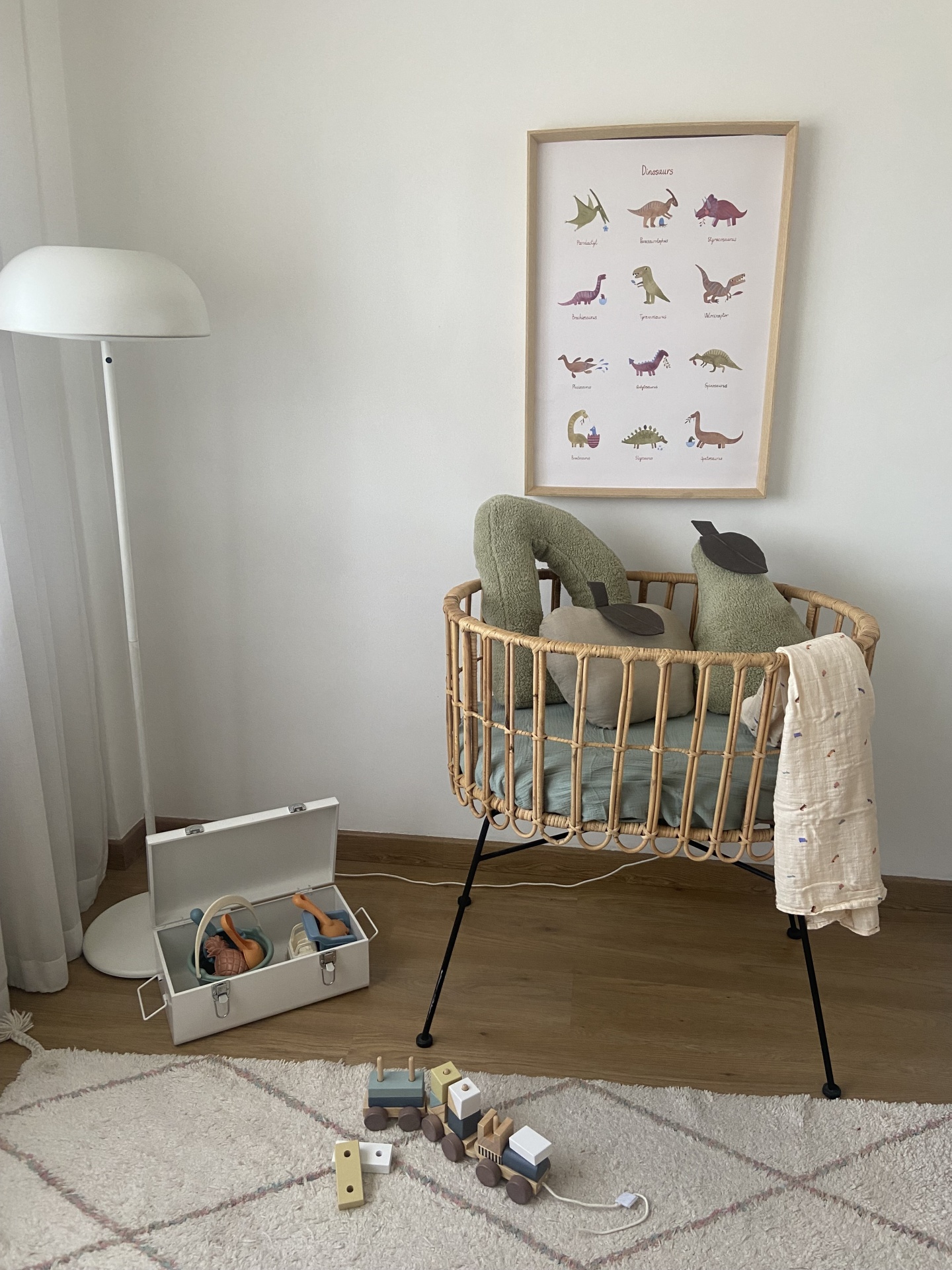 Berço de bebé rattan com almofadas verdes, candeeiro branco, brinquedos e quadro de dinossauros
