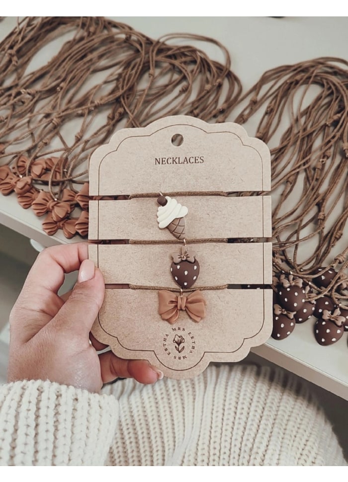 Três colares com pendentes de silicone em formas de gelado, morango e fita, em cartão reciclado com texto NECKLACES.