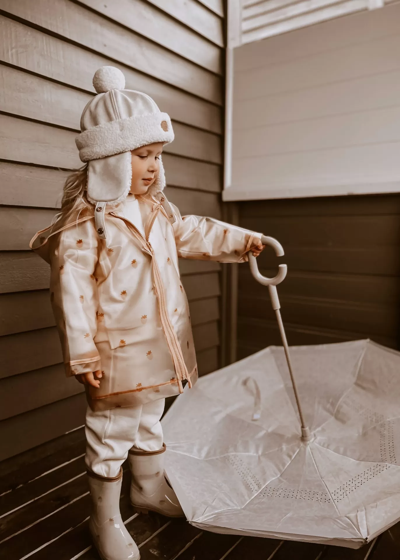Menina com casaco bege segurando guarda-chuva branco aberto junto a parede de madeira