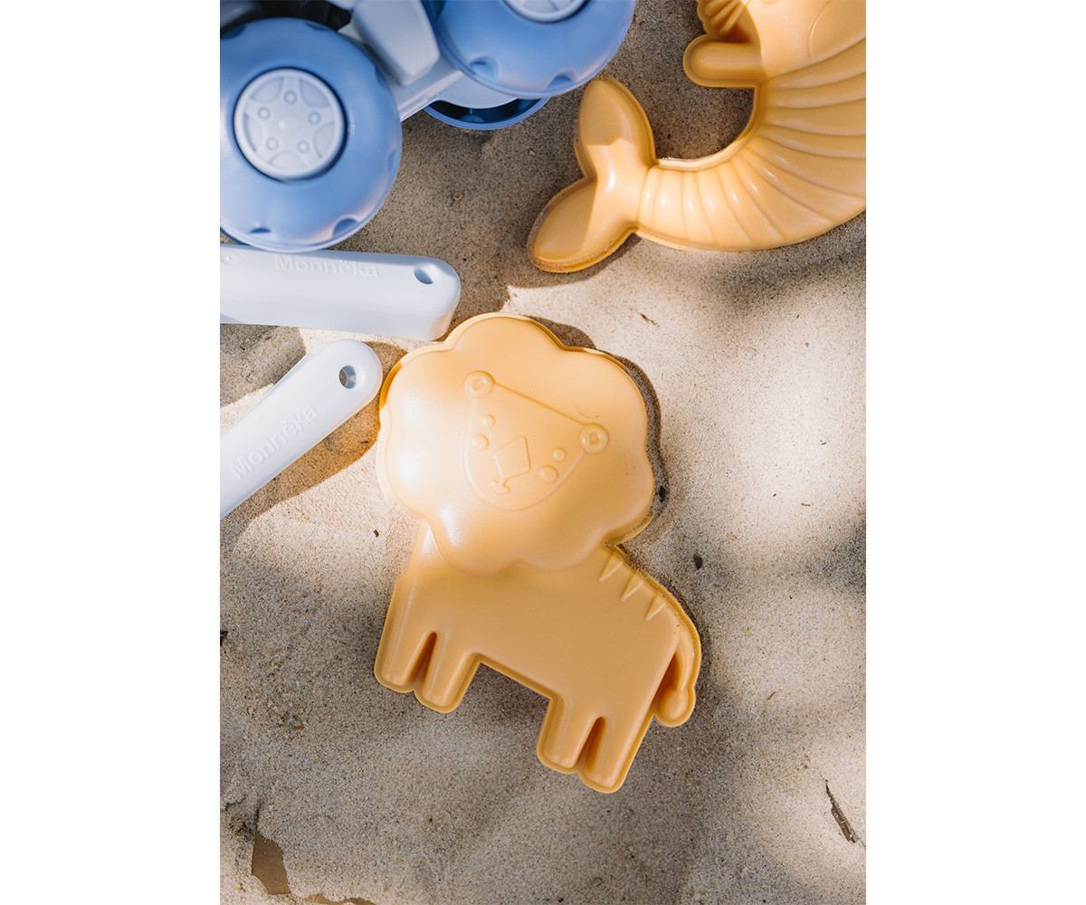 Brinquedos de areia em forma de leão e peixe, com pegas brancas gravadas com Monnéka, sobre areia.