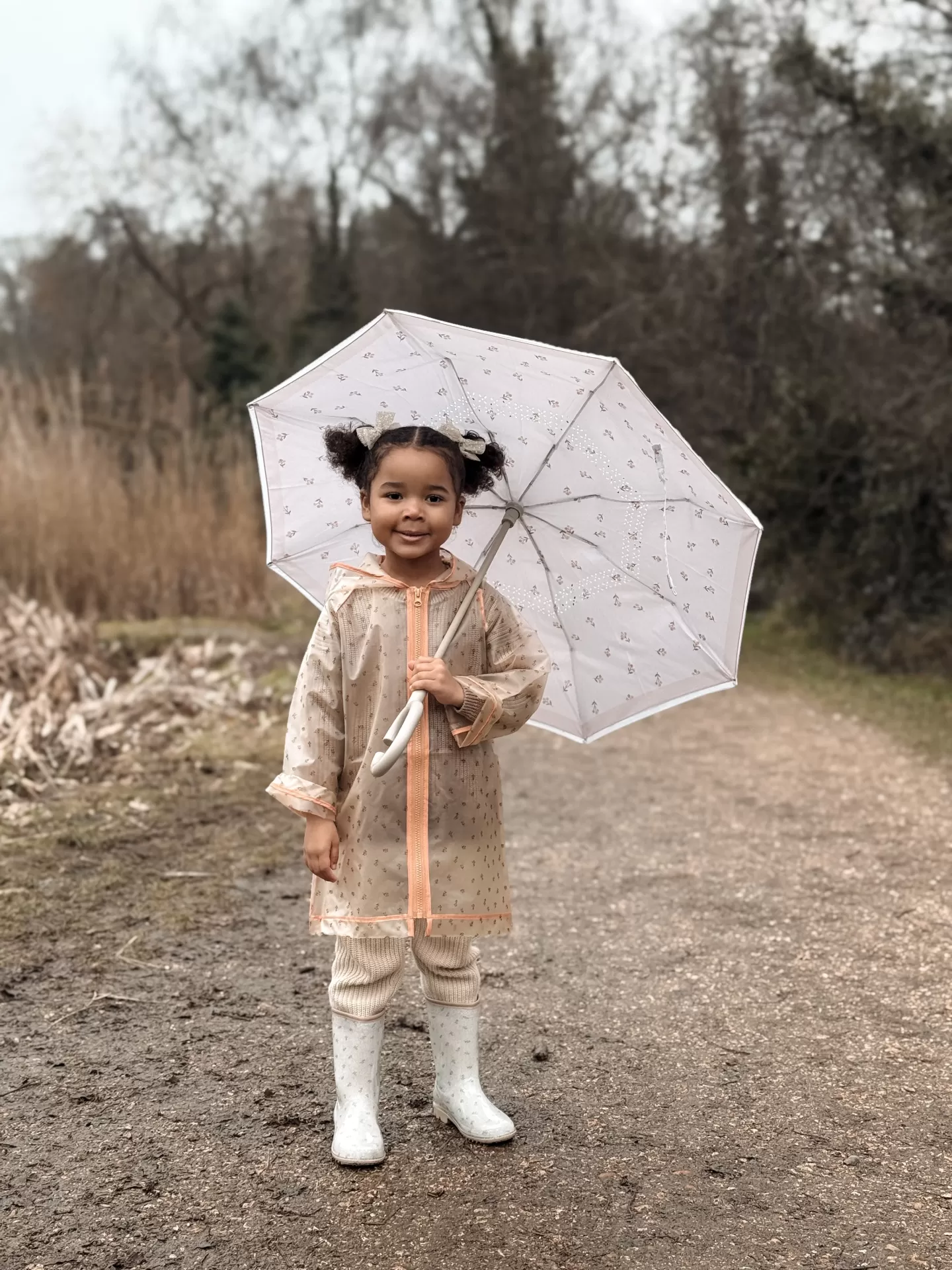 Criança com casaco impermeável bege, botas brancas e guarda-chuva branco numa trilha de terra
