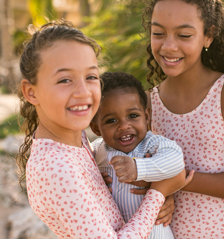 Duas crianças com roupa de banho rosa de manchas vermelhas seguram bebé com pijama riscado azul e branco ao ar livre