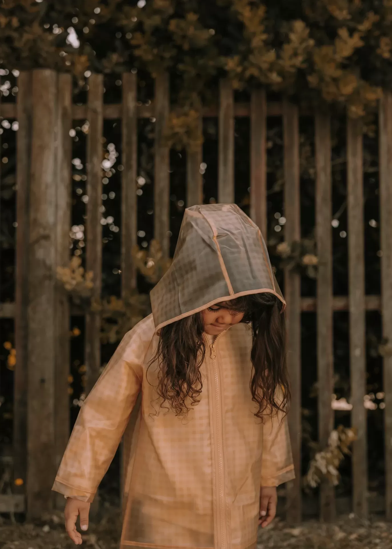 Criança com casaco impermeável bege e capuz cinza junto a uma cerca de madeira