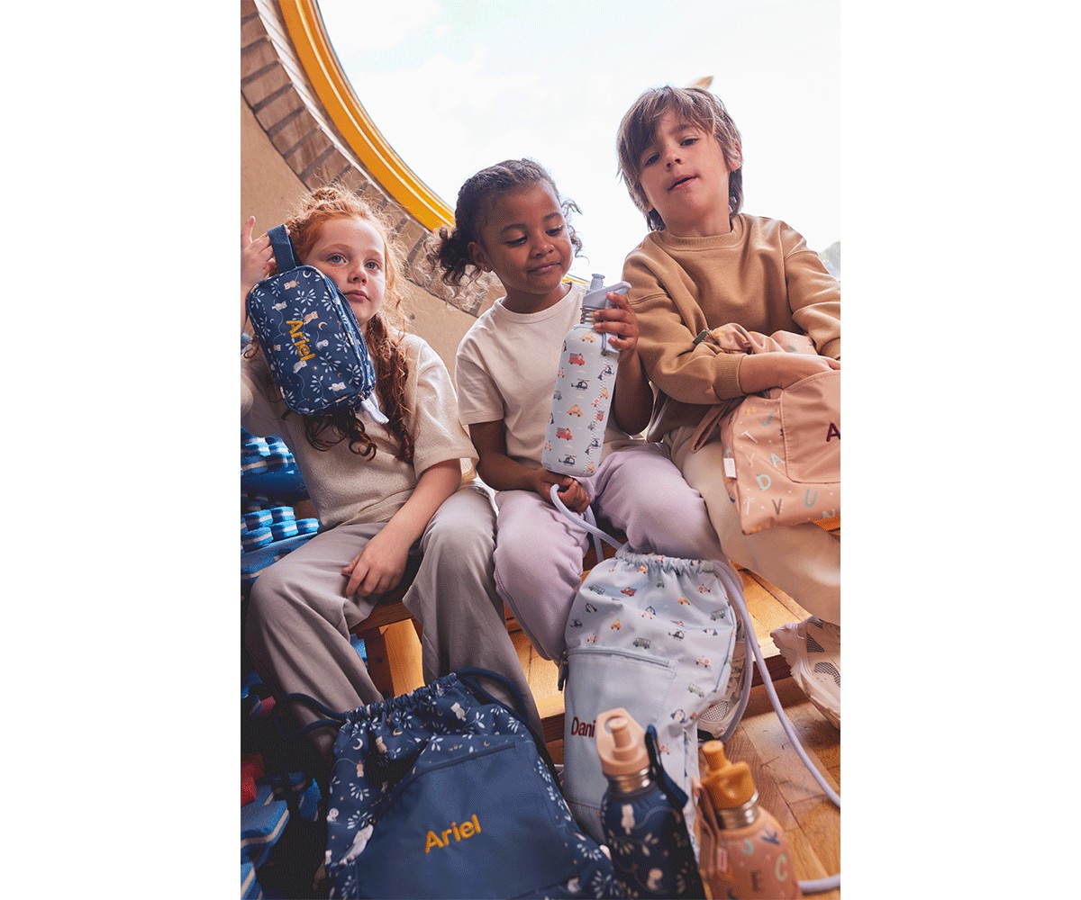 Três crianças com mochilas coloridas e garrafas reutilizáveis sentadas em ambiente luminoso