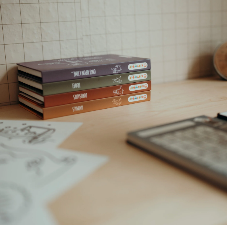 Quatro livros coloridos empilhados numa mesa clara com azulejos brancos ao fundo.