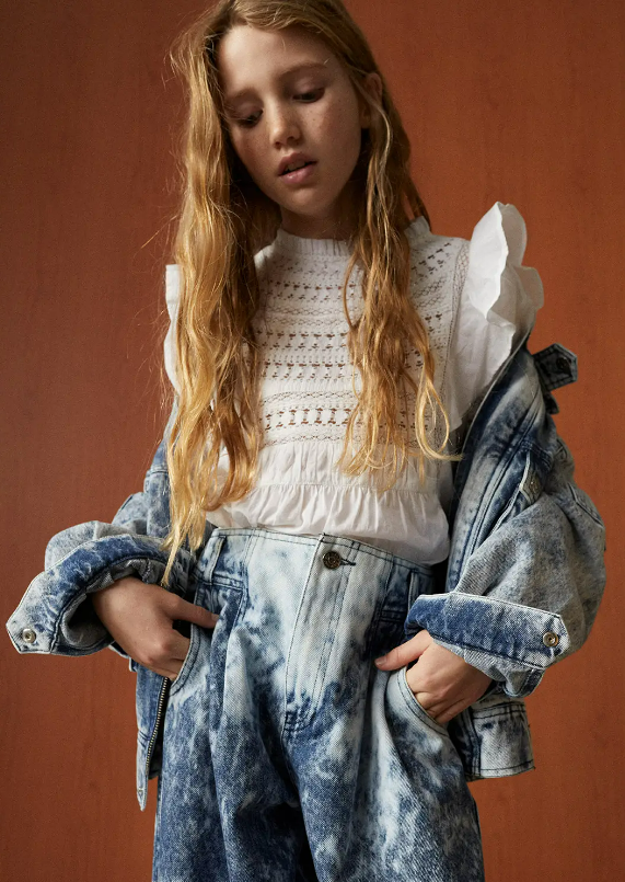 Menina com blusa branca de malha, jaqueta e calças de ganga azuis em fundo castanho