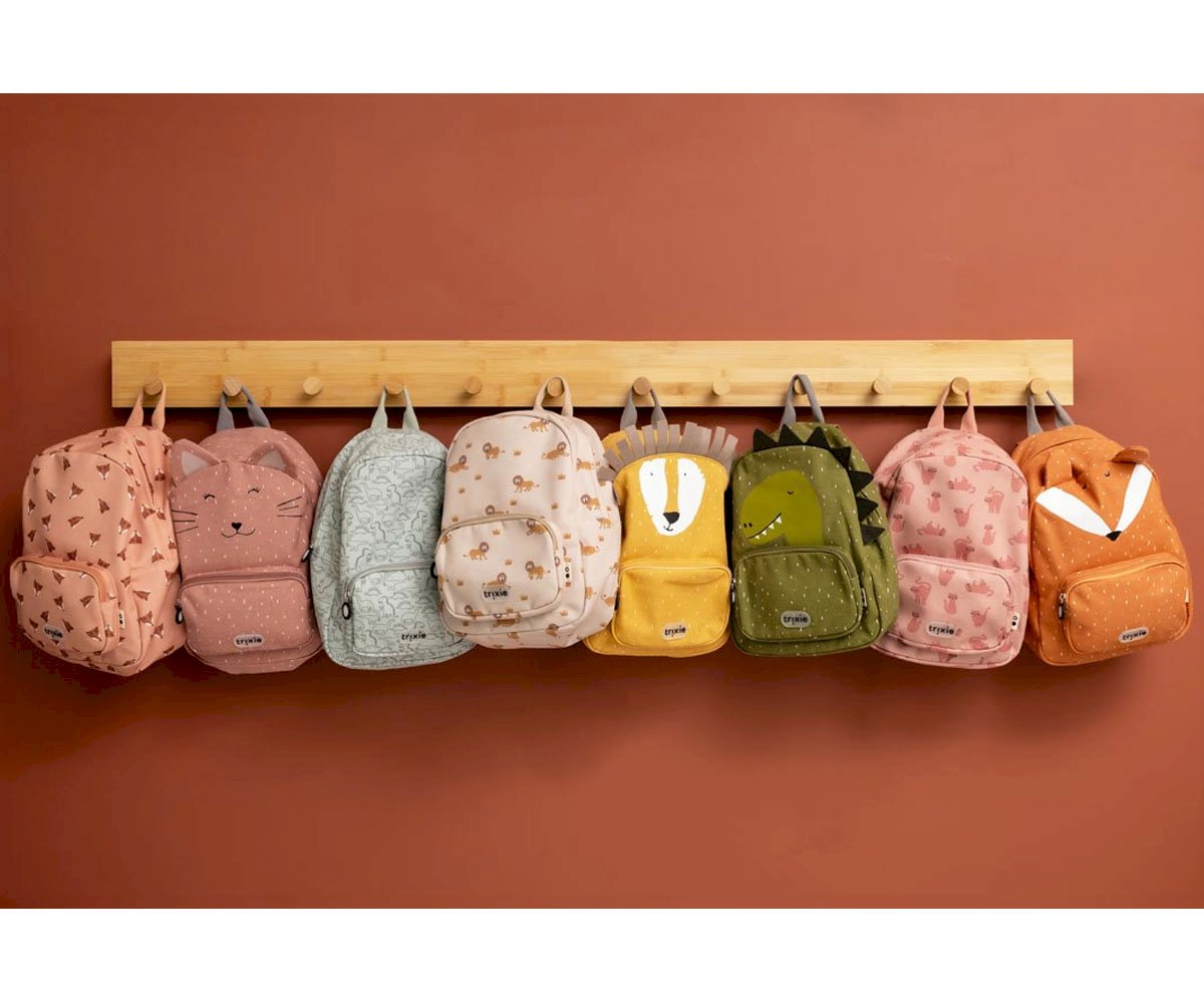 Sete mochilas infantis coloridas penduradas numa barra de madeira numa parede cor de tijolo