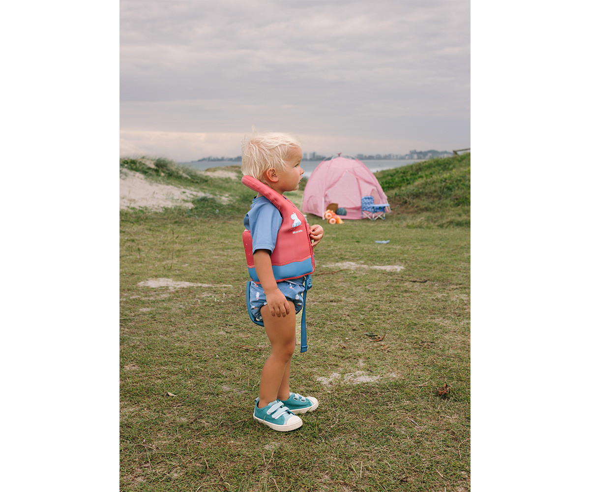Criança com colete salva-vidas rosa e azul, calças de banho e ténis azuis em relvado com tendas cor-de-rosa ao fundo