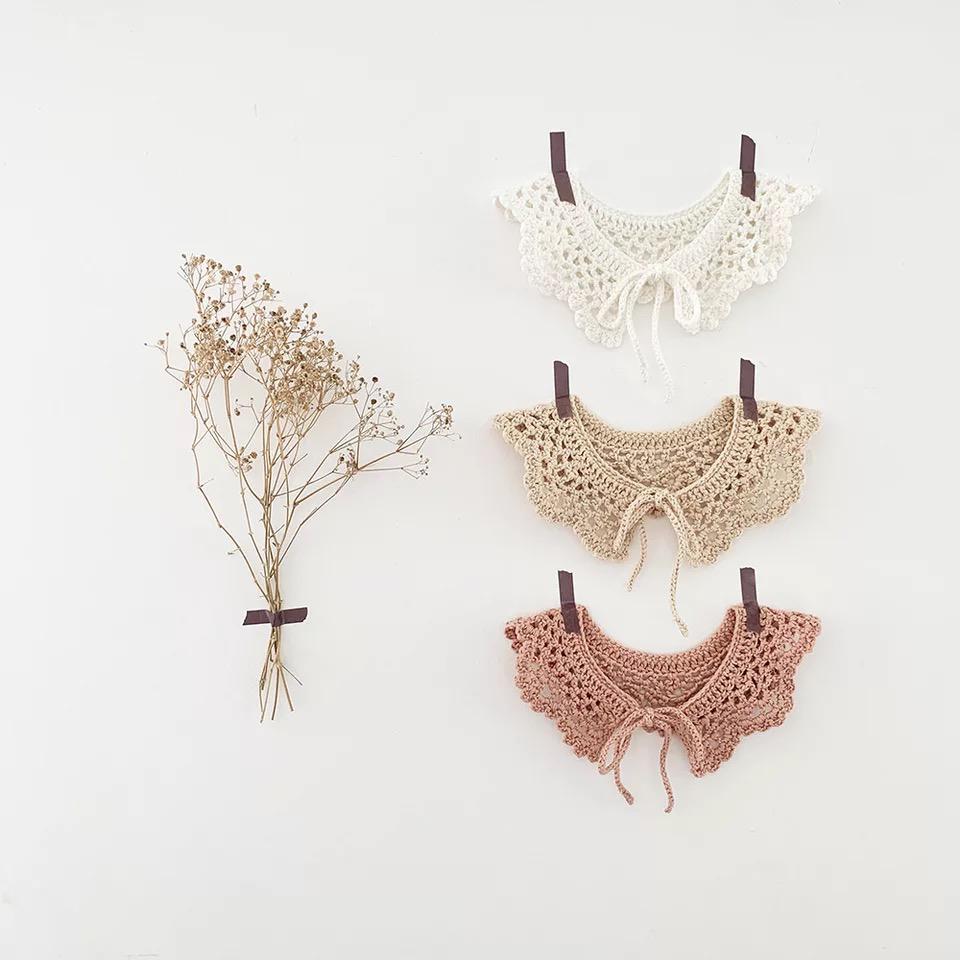 Três golas de croché em creme, bege e castanho claro penduradas na parede, ao lado um ramo seco.