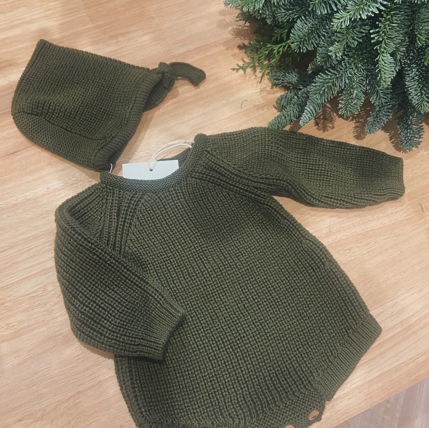 Vestido de malha verde-oliva para bebé com gorro e ramos de pinheiro ao lado