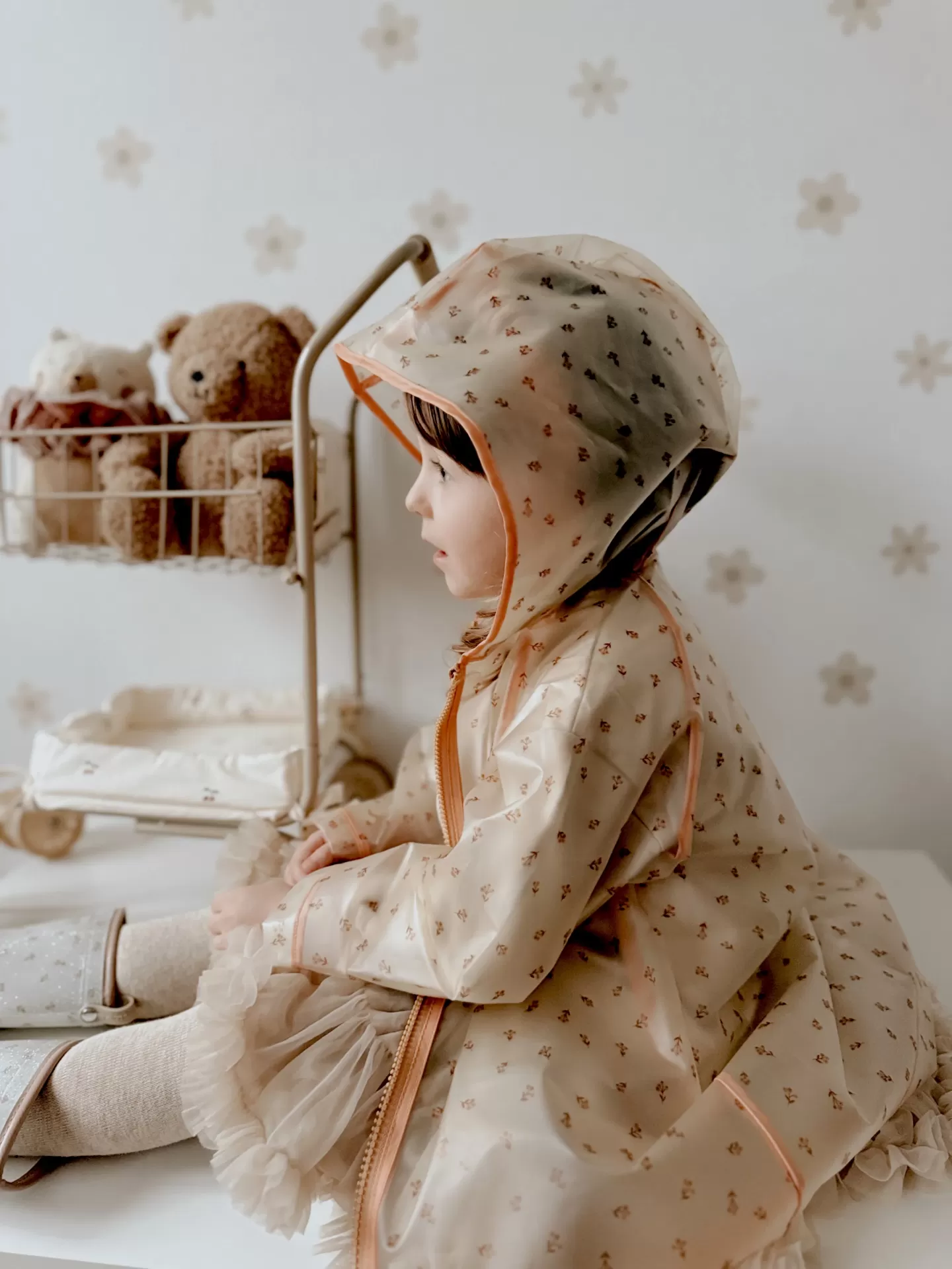 Criança com impermeável bege de padrão floral com capuz, sentada, com brinquedos atrás