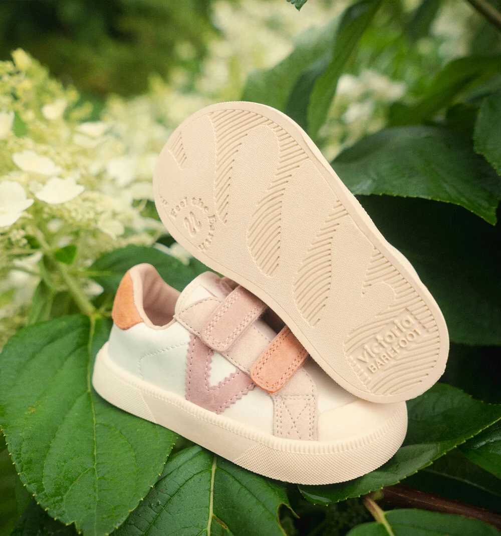 Sapatilhas infantis Victoria Barefoot em pele branca com detalhes em camurça rosa e castanho claro sobre folhas verdes.