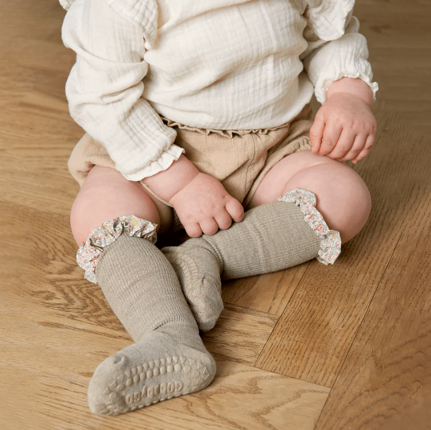 Bebé sentado com camisa branca, calças bege e meias cinza com babados florais