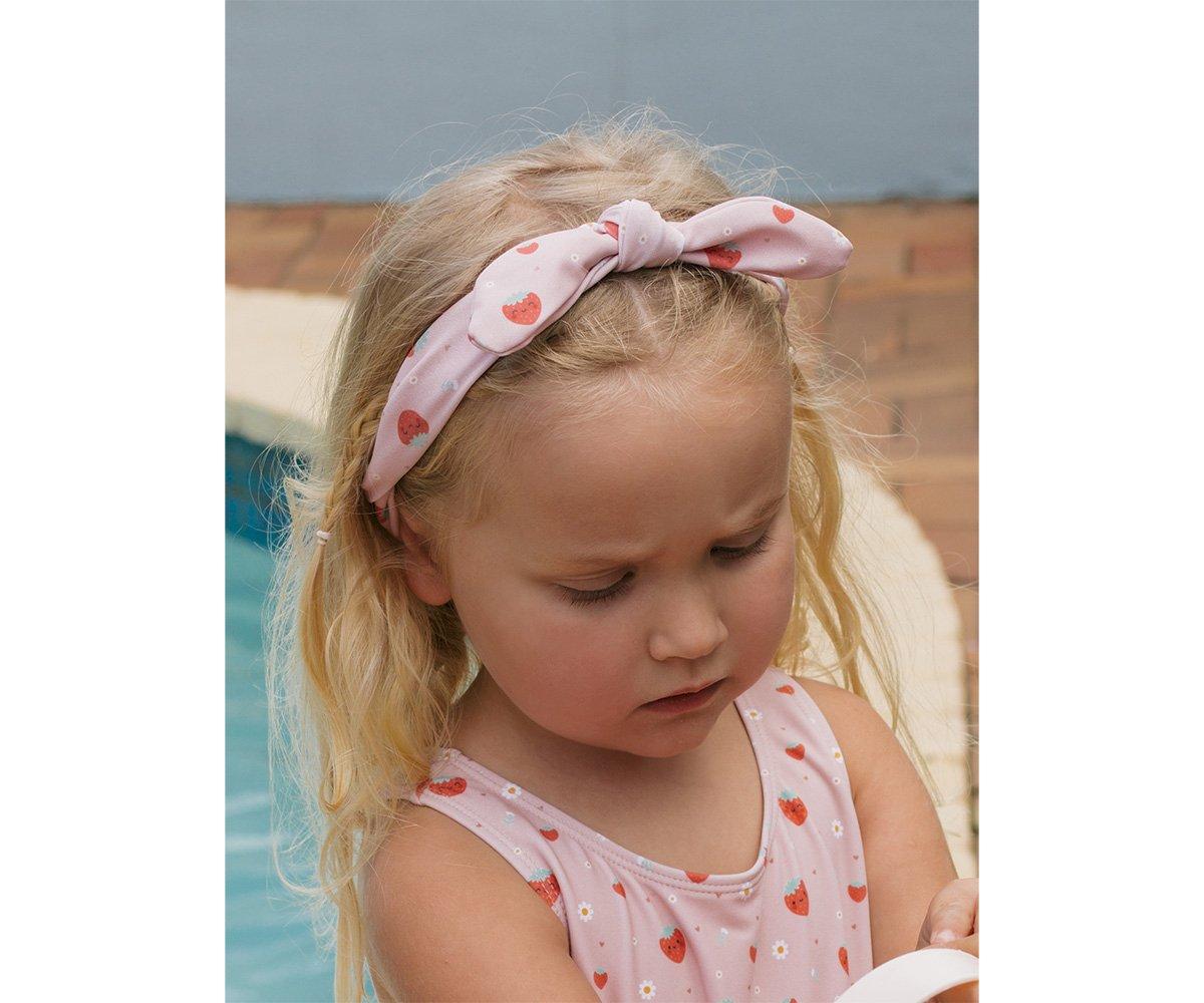 Criança loira com faixa de cabelo rosa claro e vestido com estampas vermelhas ao lado de piscina