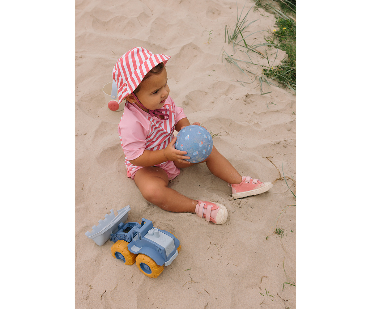 Bebé com chapéu rosa e branco, roupa rosa e estiver sentado na areia a brincar com bola azul e brinquedo escavadora.