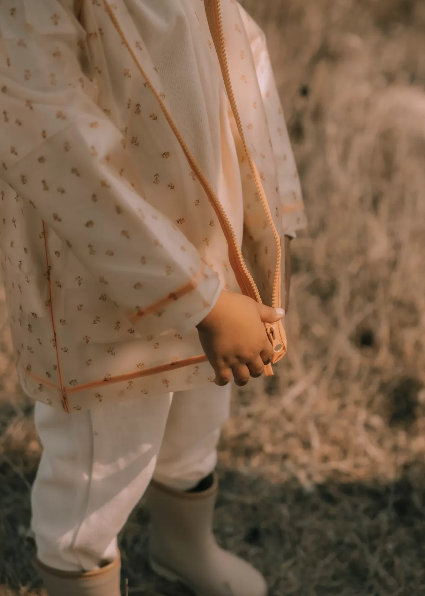 Criança com casaco bege estampado, calças brancas e botas bege ao ar livre