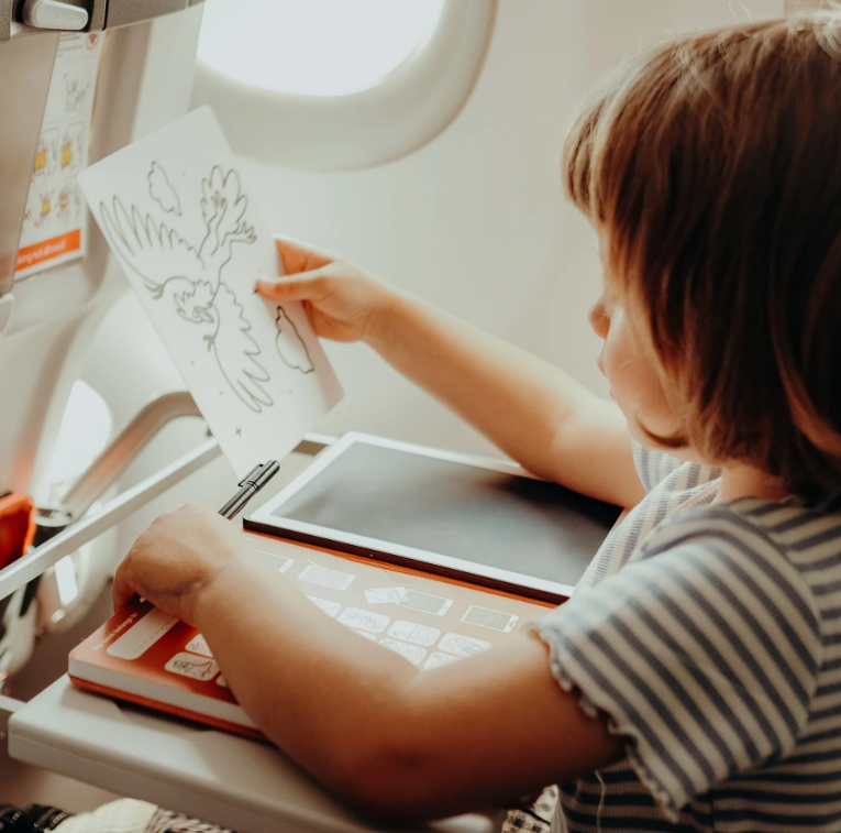 Criança com desenho para colorir, tablet e livro numa bandeja de avião