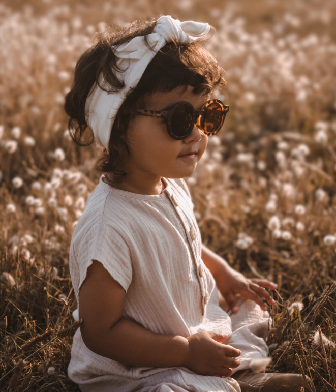 Criança com óculos de sol e faixa de cabelo branca sentada num campo de flores secas.