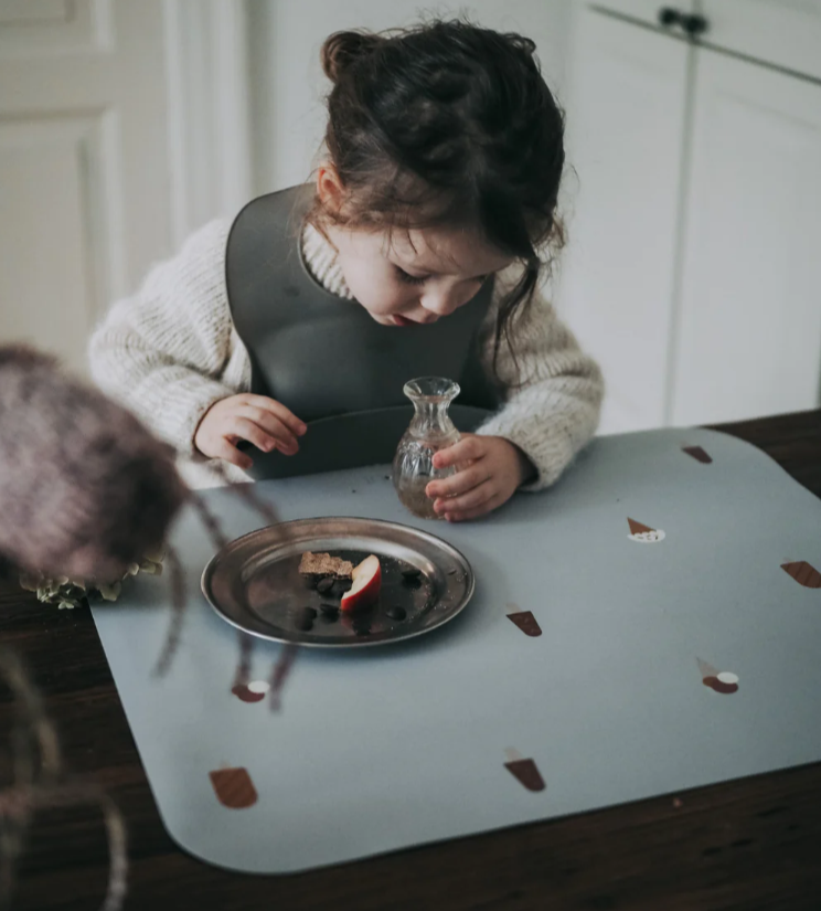 Bebé com babete cinzento a segurar jarro de vidro junto a prato metálico com comida numa mesa com base azul clara decorada