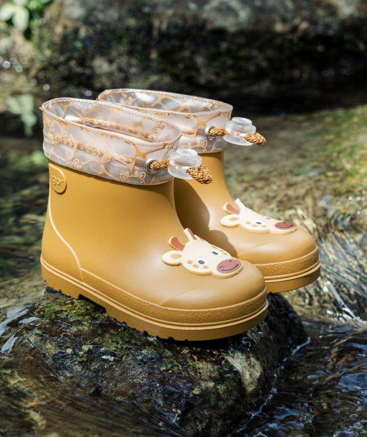 Botas de chuva infantis amarelas com estampa de vaca e detalhe transparente decorado, sobre pedra junto à água.