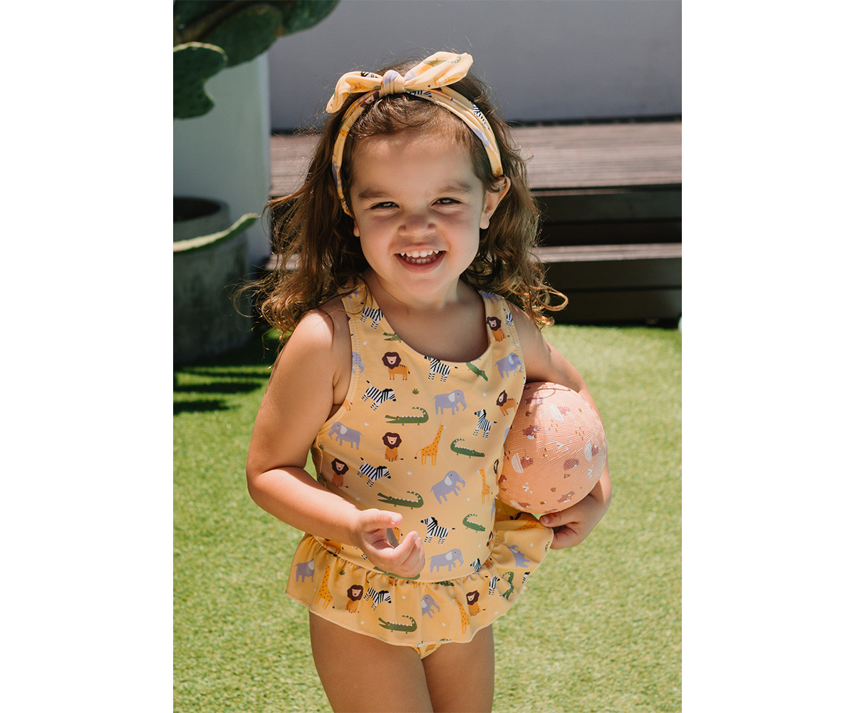 Menina com fato de banho amarelo com estampado de animais e faixa de cabelo, segura bola cor-de-rosa no relvado.