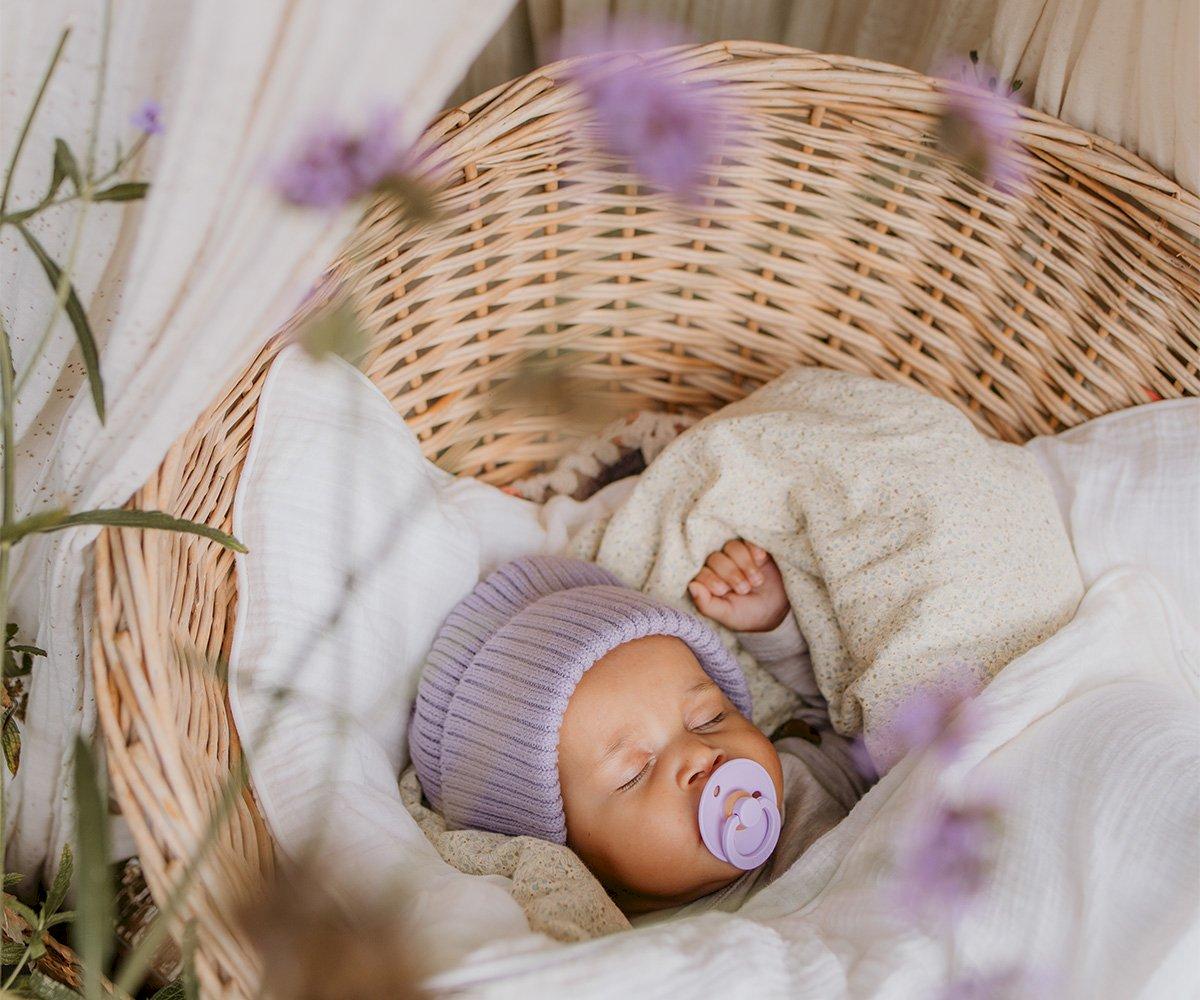 Bebé a dormir num berço de verga com chupeta lilás e gorro roxo