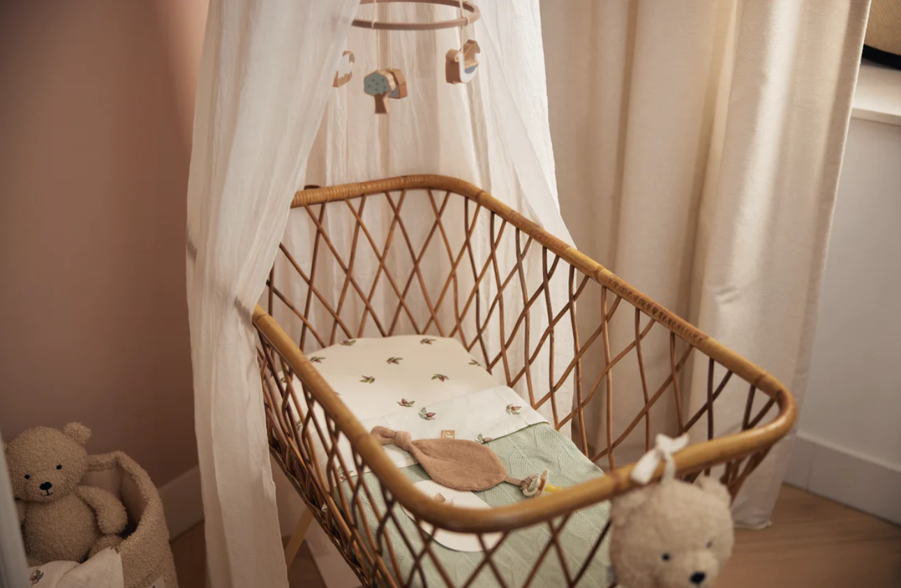 Berço de bebé em vime com almofada branca e mosquiteiro branco num quarto com ursinho de peluche e cortinas bege