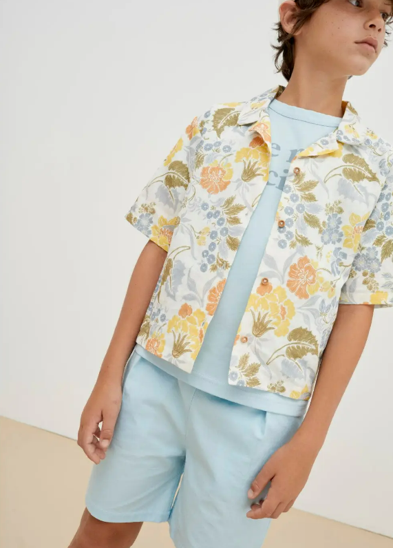 Conjunto infantil com camisa floral, t-shirt e calções azuis, cenário interior