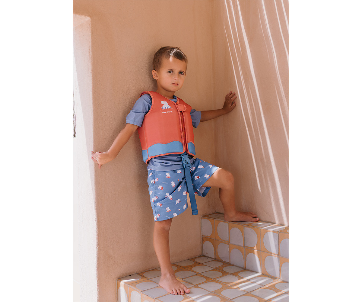 Criança de colete salva-vidas vermelho e azul com calções azuis numa escadaria de azulejos