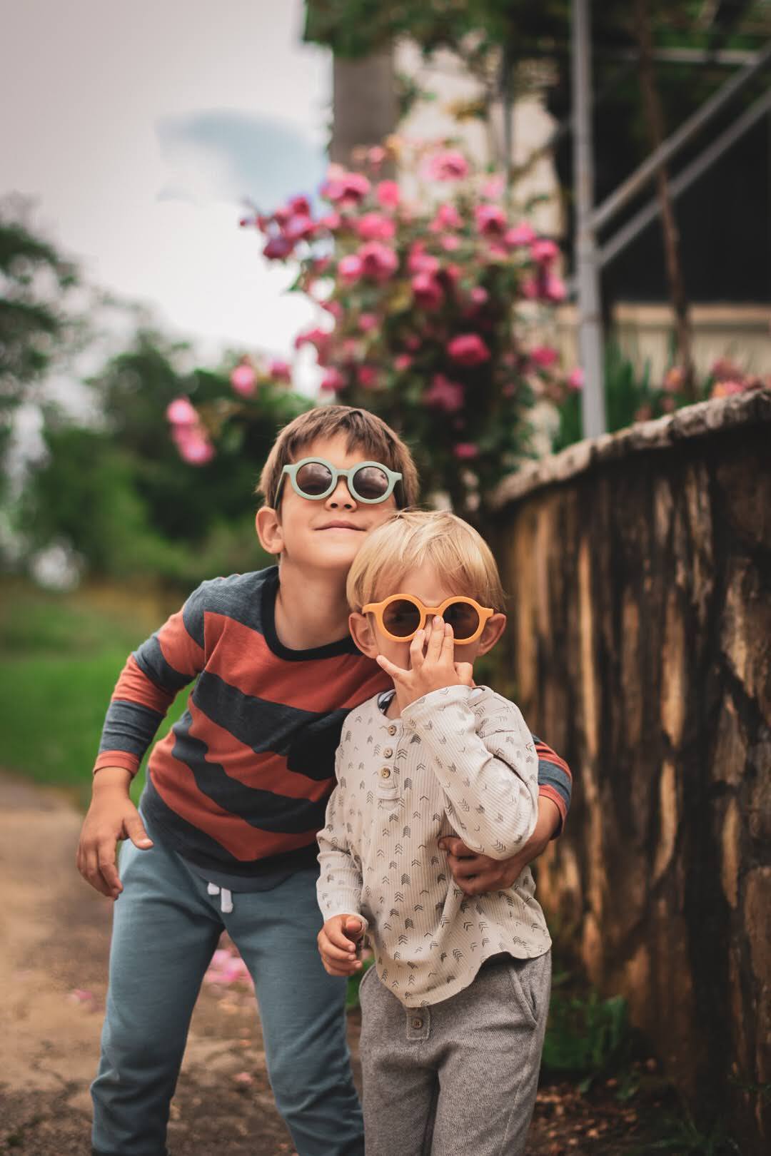 Dois meninos usando óculos de sol e roupa casual junto a um muro de pedra com flores rosas ao fundo