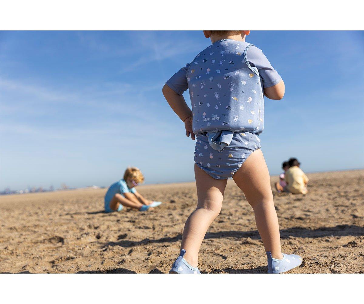 Três crianças na praia com colete salva-vidas azul claro e roupa de banho, areia e céu azul.