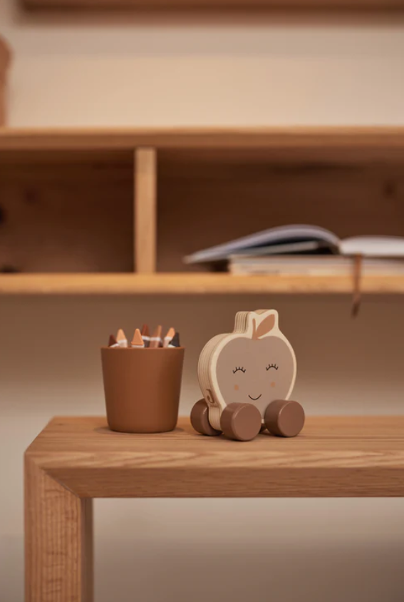 Lápis de cor em copo castanho e brinquedo de madeira em forma de maçã sorridente, sobre mesa de madeira clara.