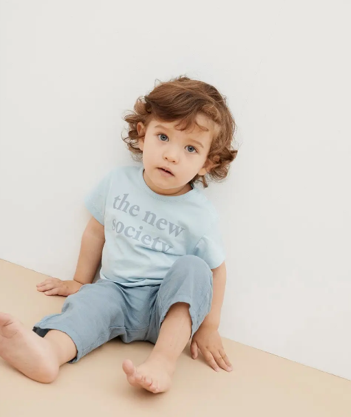 Criança sentada no chão com camiseta azul clara e calças azuis, texto 'the new society' na camiseta