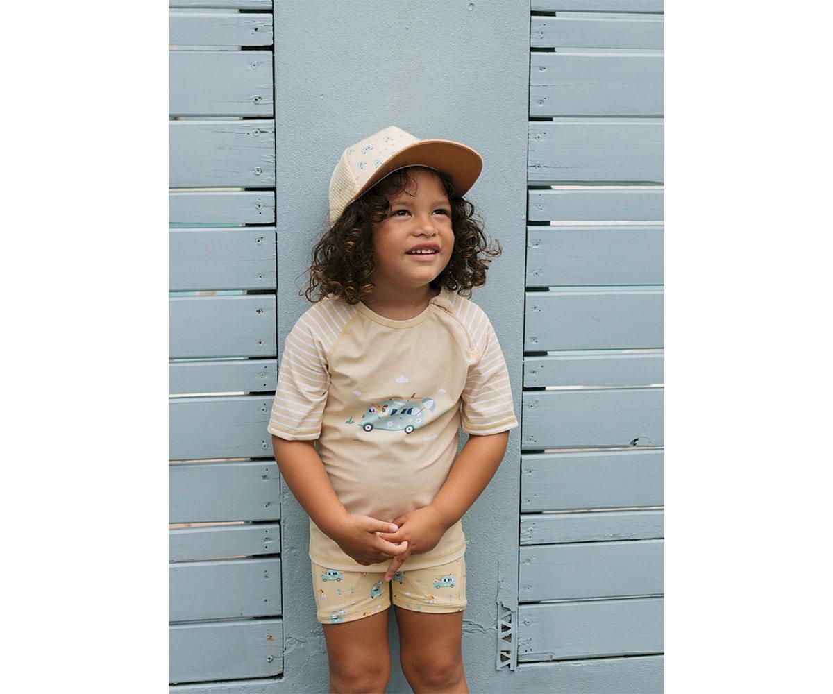 Menino com roupa creme e chapéu bege em frente a parede cinza claro