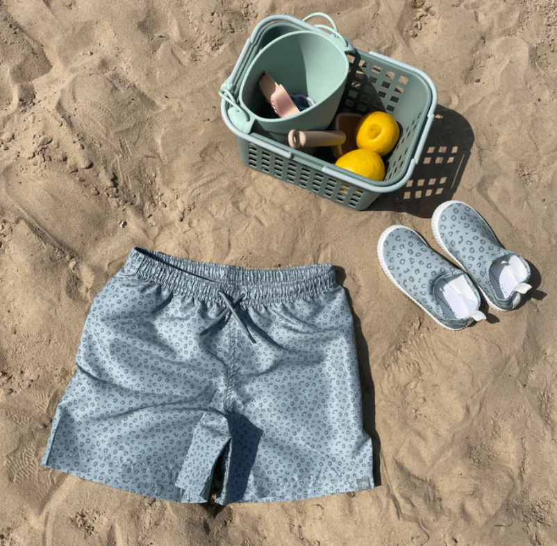 Calções de banho azuis com padrão de pequenas flores e sapatilhas de água cinzentas na areia junto a brinquedos de praia