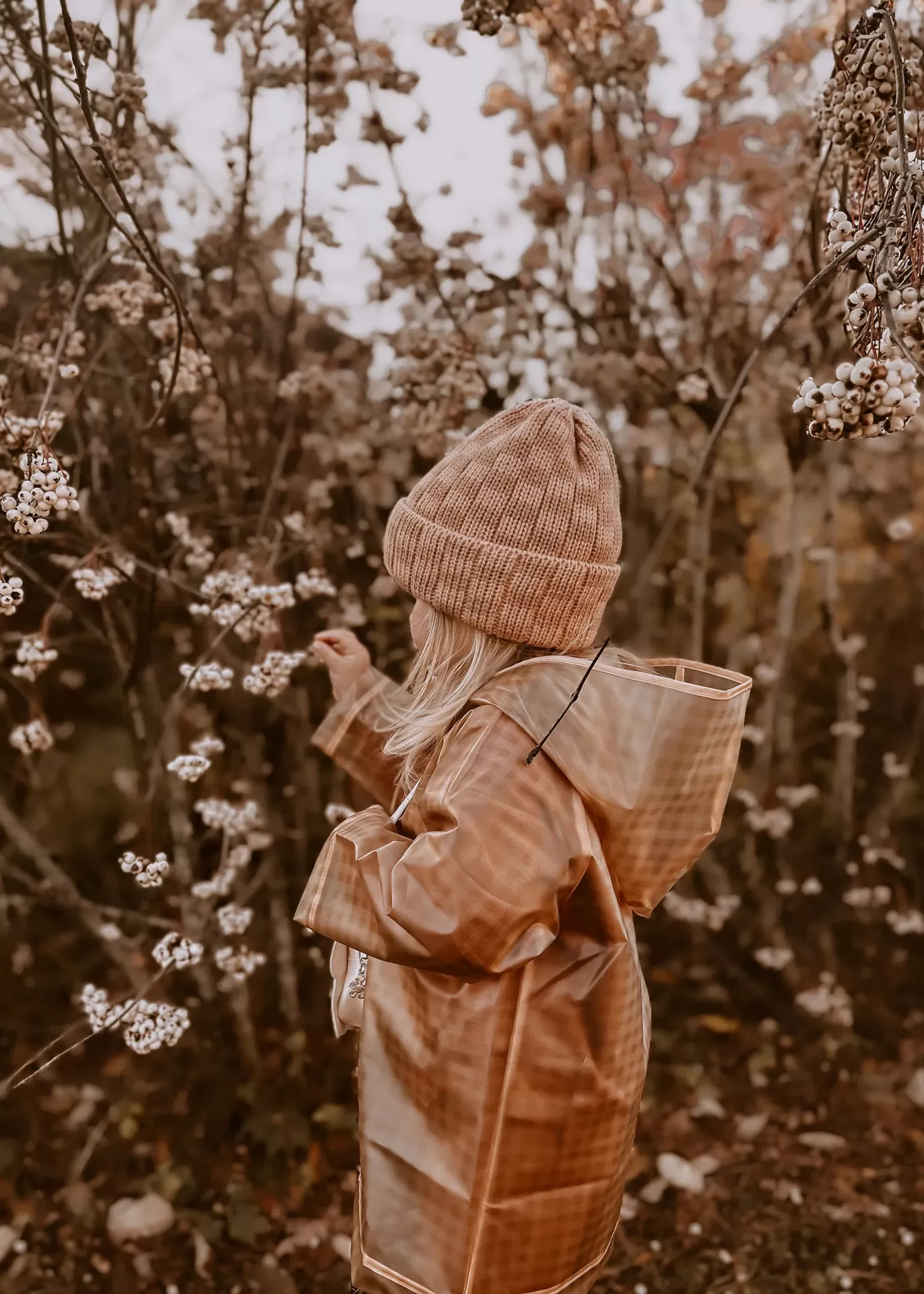 Criança com capa de chuva castanha e gorro de malha em ambiente natural outonal