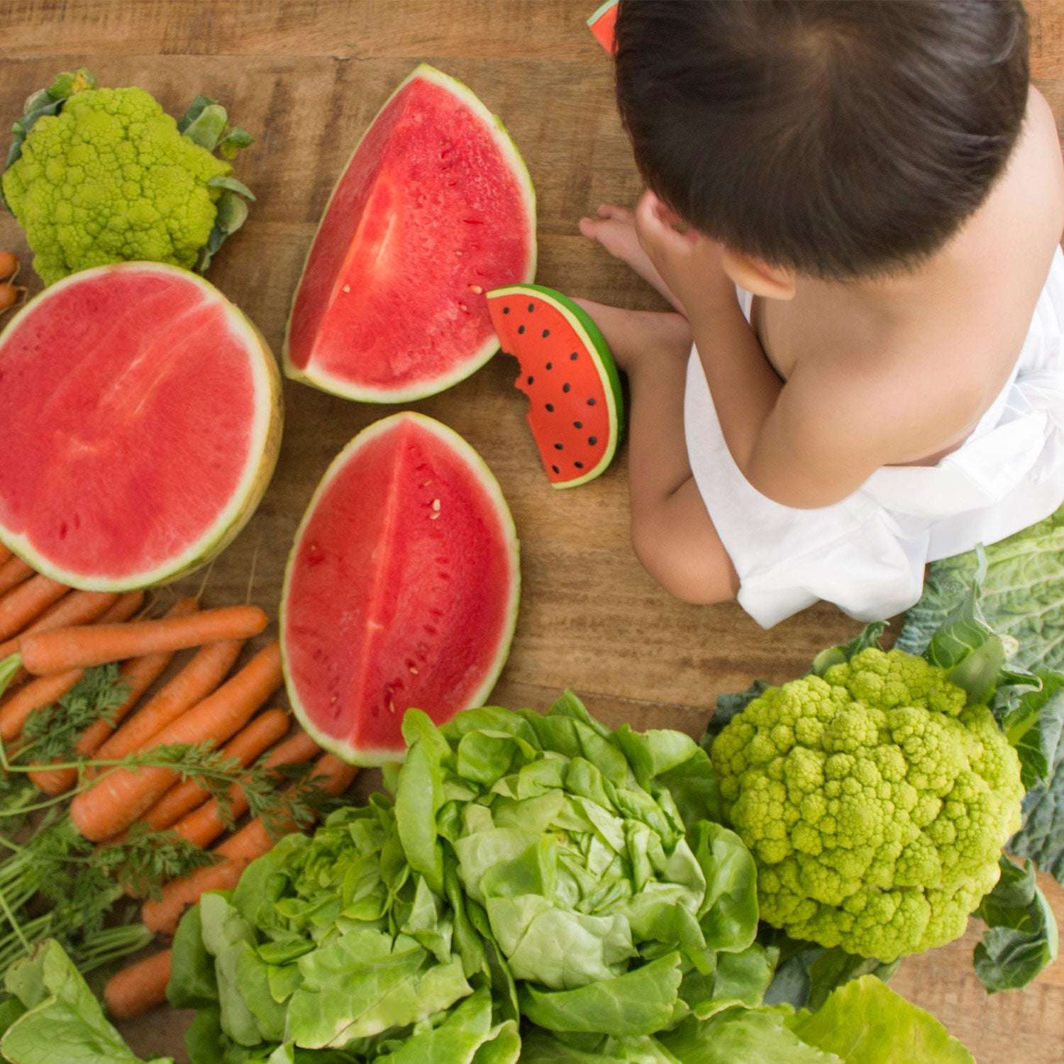 Criança sentada ao lado de melancias cortadas e vários vegetais frescos sobre madeira