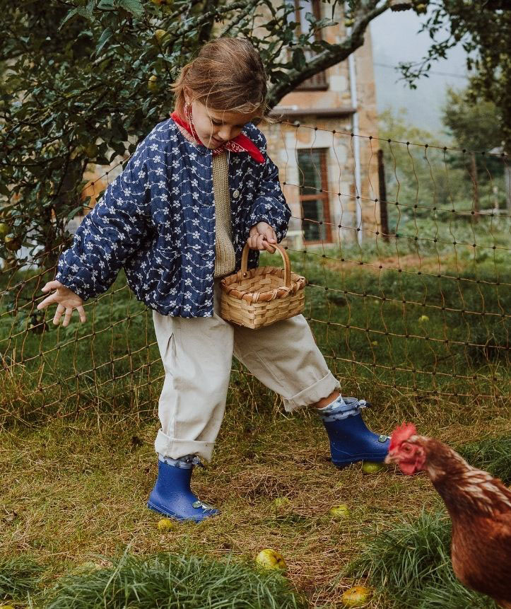 Criança com casaco azul e calças bege segurando um cesto de verga em ambiente rural com galinha