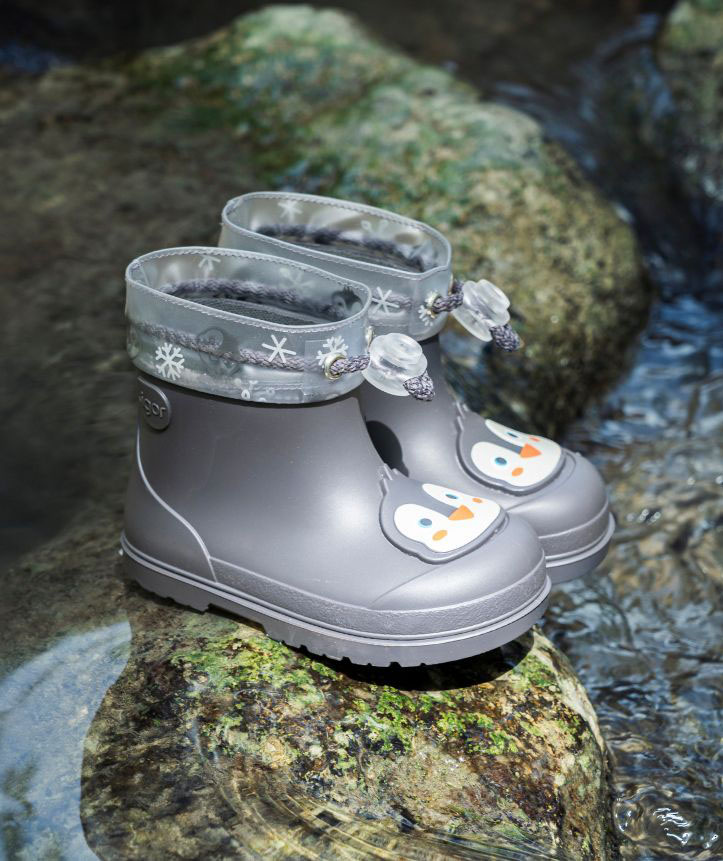 Botas de borracha cinzentas infantis com pinguins e detalhes de flocos de neve em ambiente com pedra e água.