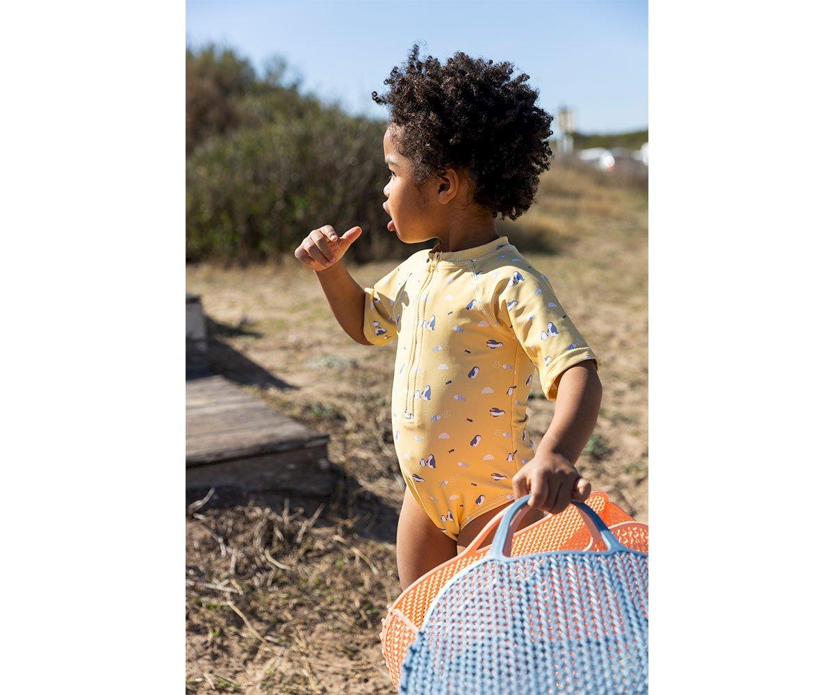 Criança em fato de banho amarelo segurando mochila trançada em ambiente exterior