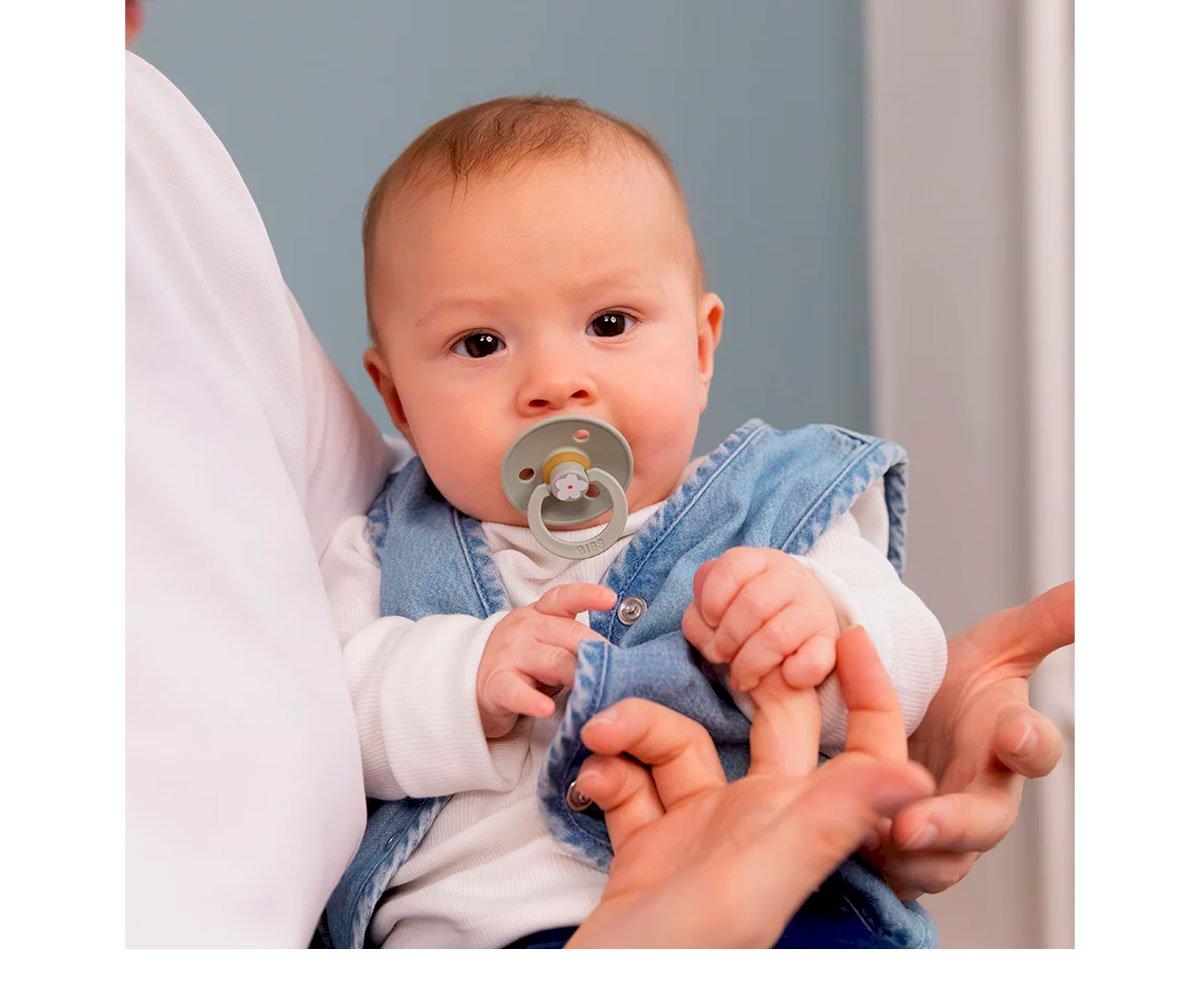 Bebé com chupeta e roupa azul jeans e branca, segurando cordão da chupeta